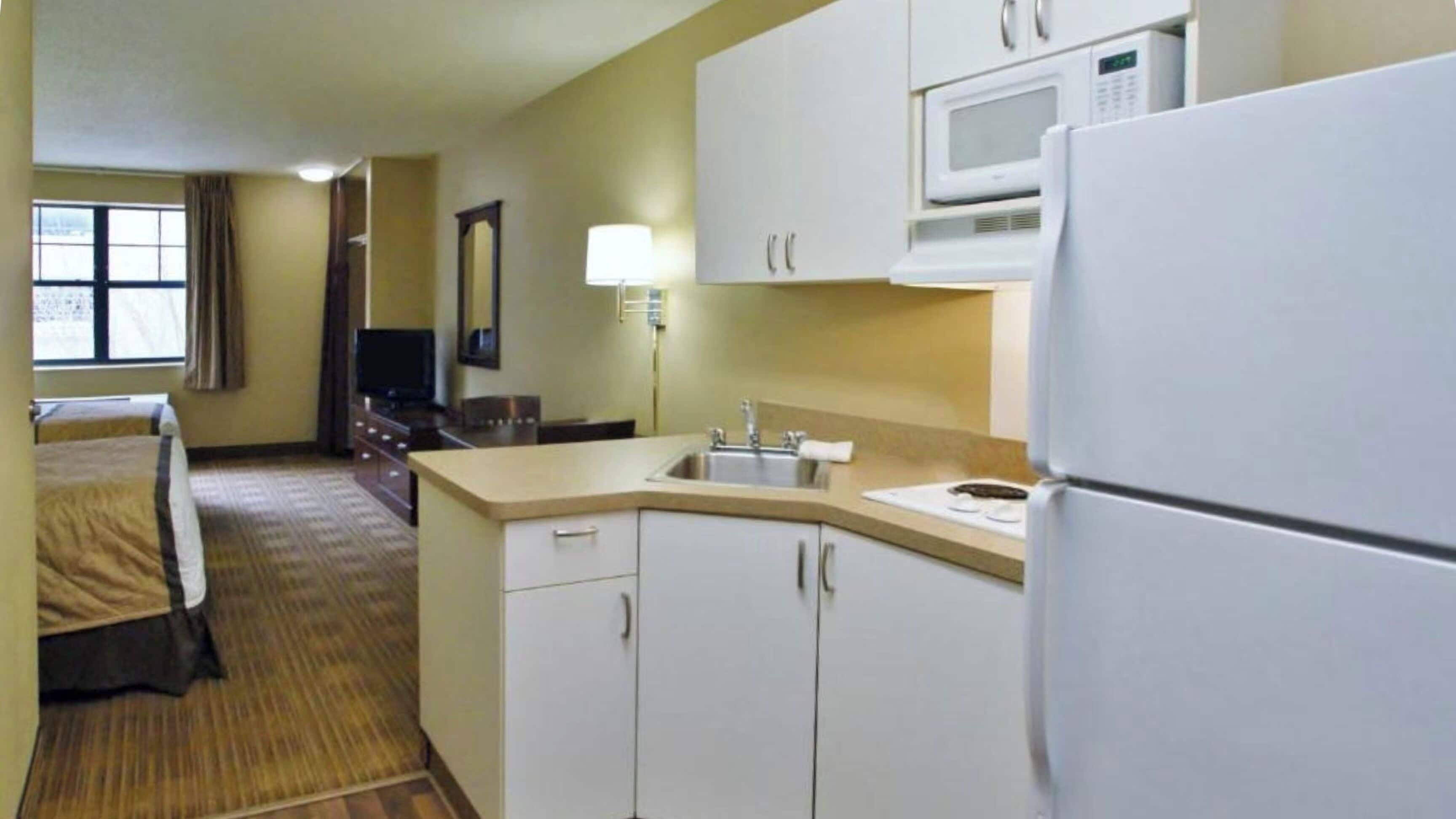 A compact hotel room with a kitchenette, a bed, a television, and a window with curtains.