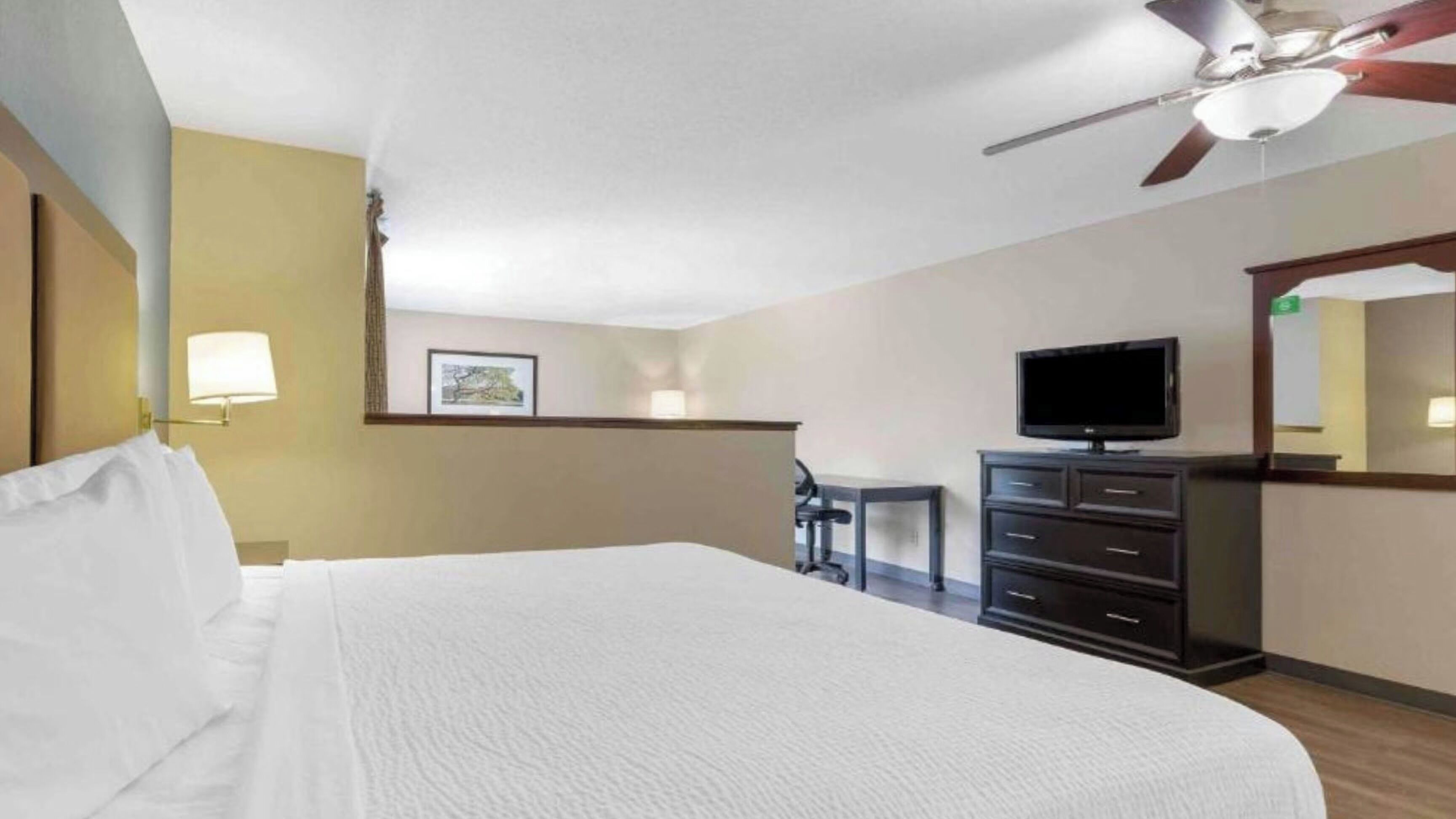 A hotel room with a bed, a desk with a chair, a television, and a ceiling fan.