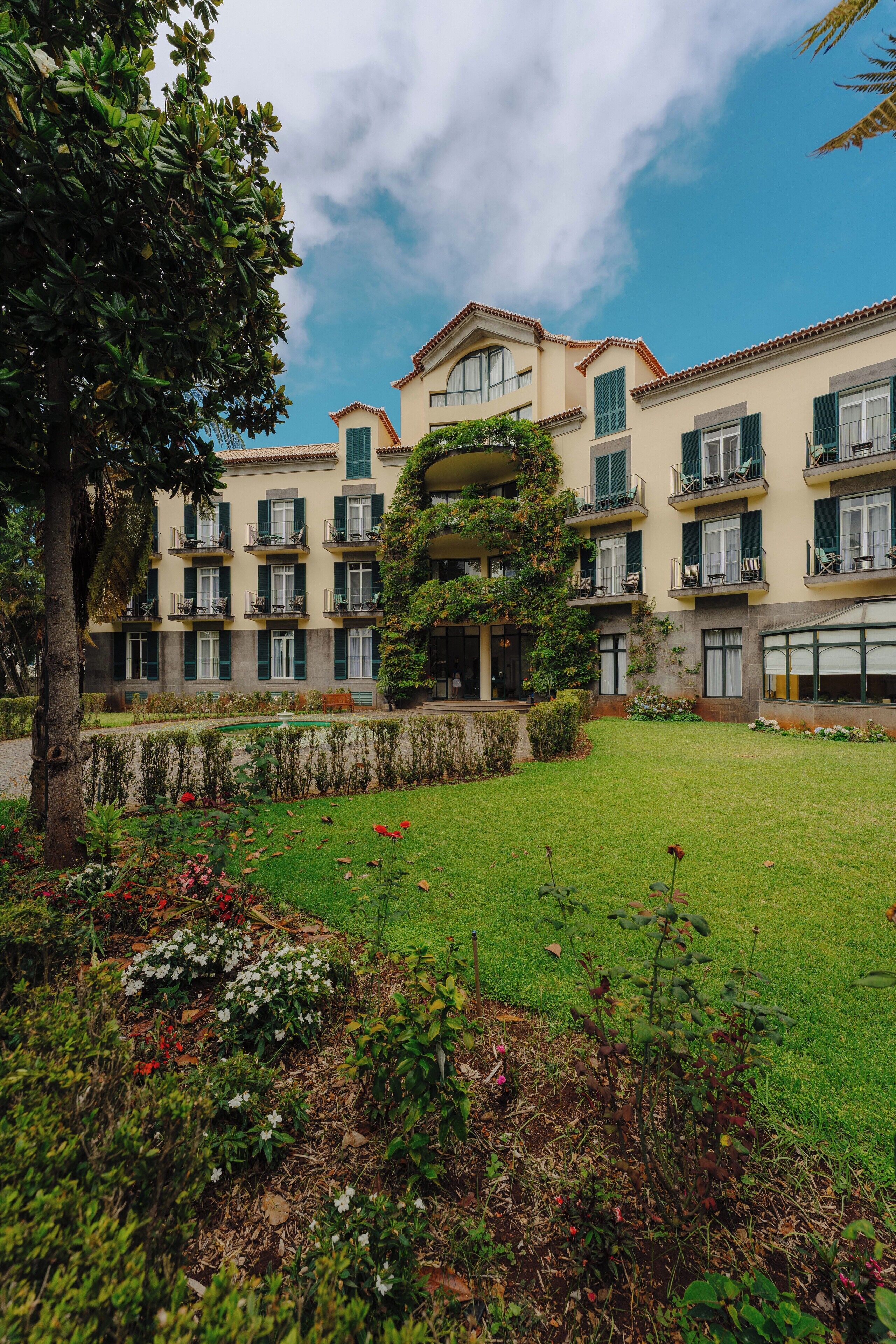 Un hotel de varios pisos con balcones, un césped cuidado y un jardín con diversas plantas y flores.