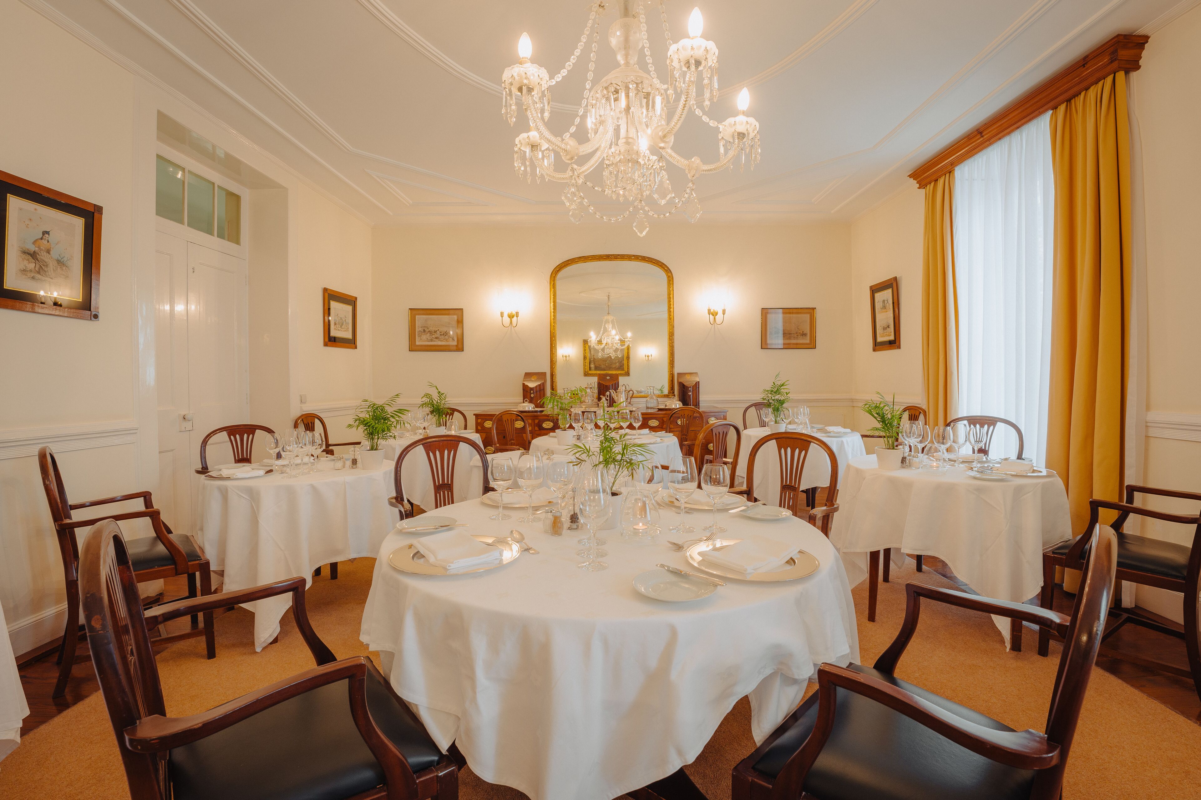 Une salle à manger élégante, avec plusieurs tables rondes, chacune recouverte d’une nappe blanche et mise avec de l’argenterie et de la vaisselle. Des chaises en bois sont disposées autour des tables, et un grand lustre est suspendu au plafond.