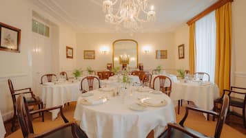 Une salle à manger élégante, avec plusieurs tables rondes, chacune recouverte d’une nappe blanche et mise avec de l’argenterie et de la vaisselle. Des chaises en bois sont disposées autour des tables, et un grand lustre est suspendu au plafond.