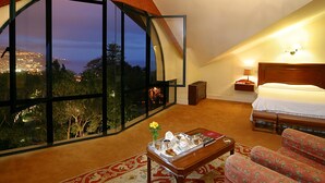 A bedroom with a large window, a bed, a nightstand, a lamp, and a view of the city at night.