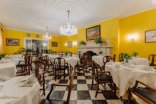 Un comedor con mesas dispuestas para el servicio, sillas, una araña y una chimenea.