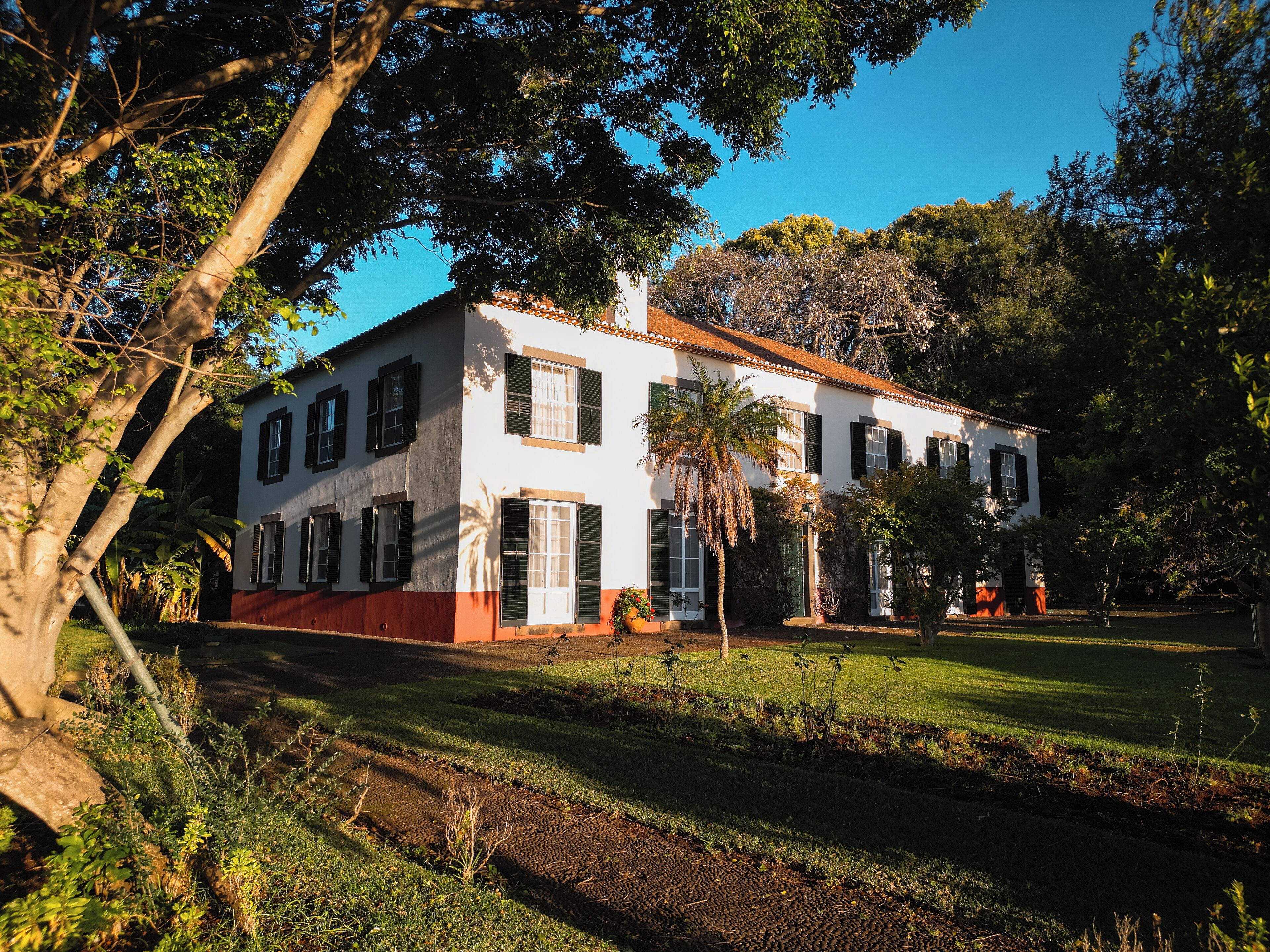 Un bâtiment blanc à deux étages, avec une base rouge, entouré d’une végétation luxuriante et d’arbres.