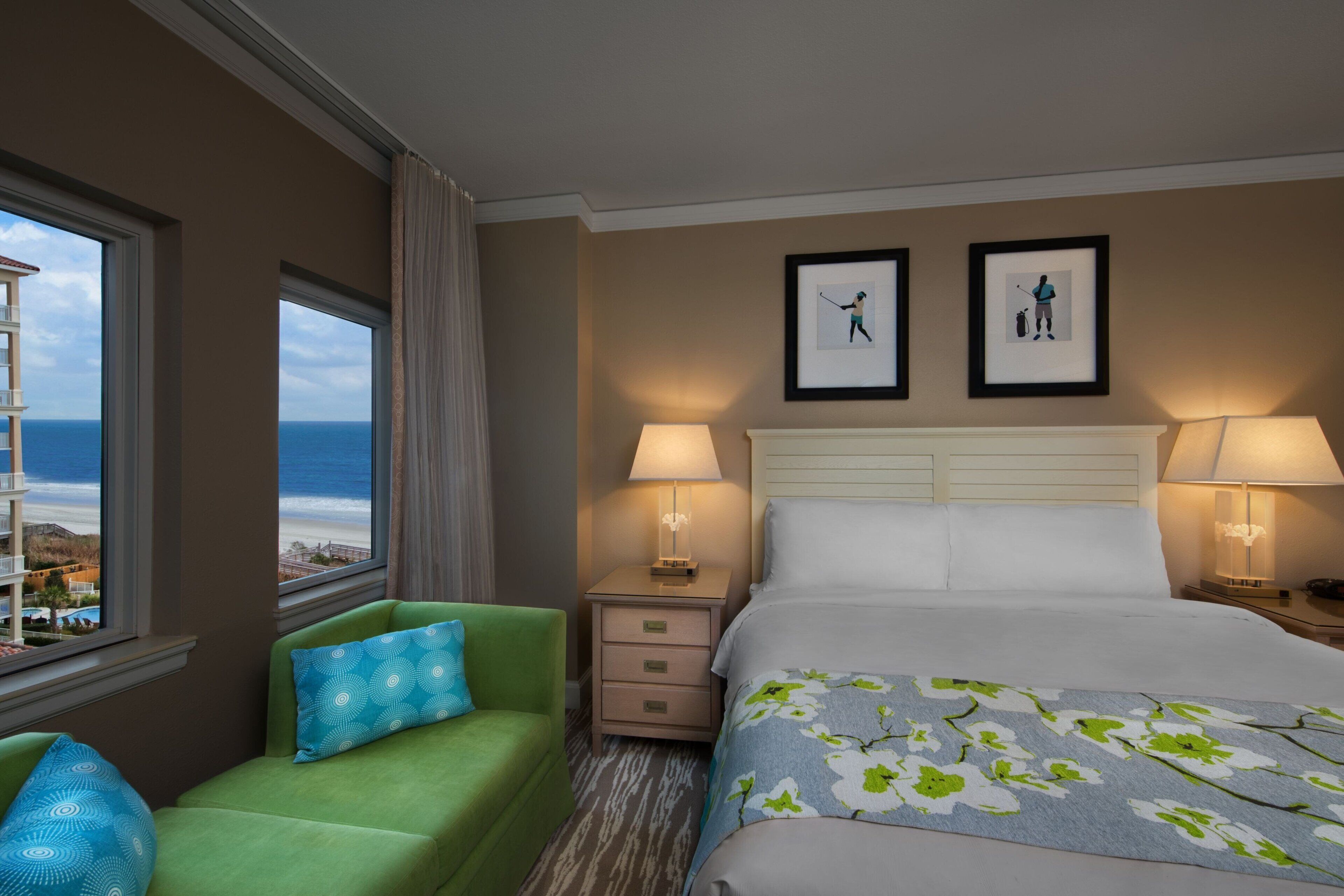 A hotel room with a bed, two bedside lamps, a nightstand, a green armchair, a window with a view of the sea, and two framed pictures on the wall.