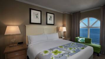 Quarto de hotel com uma cama grande, duas luminárias de cabeceira, uma poltrona verde e uma janela com vista para o oceano.