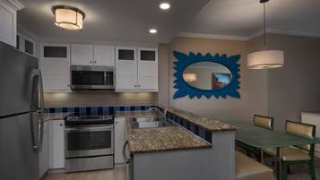 Cocina compacta con armarios blancos, un refrigerador de acero inoxidable, un microondas y una estufa. Hay una zona de comedor con una mesa y sillas.
