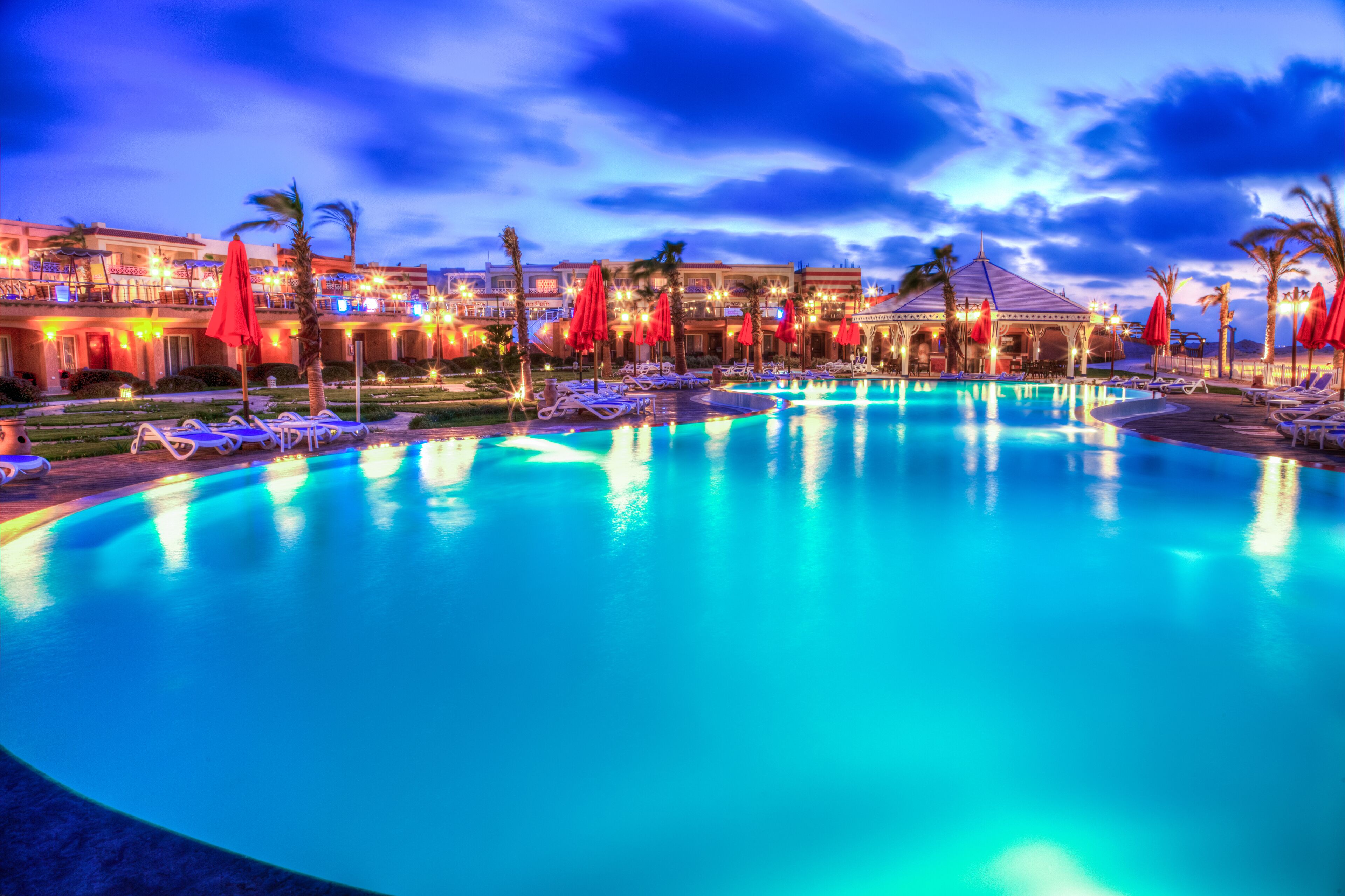 A swimming pool with lounge chairs and umbrellas, surrounded by buildings with lights.