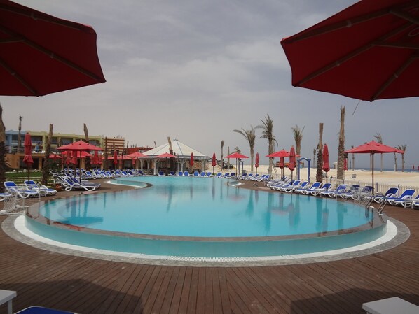 A pool area with blue lounge chairs and red umbrellas, surrounded by palm trees and a building in the background. - Porto Matrouh Beach Resort (Marsa Matrouh)