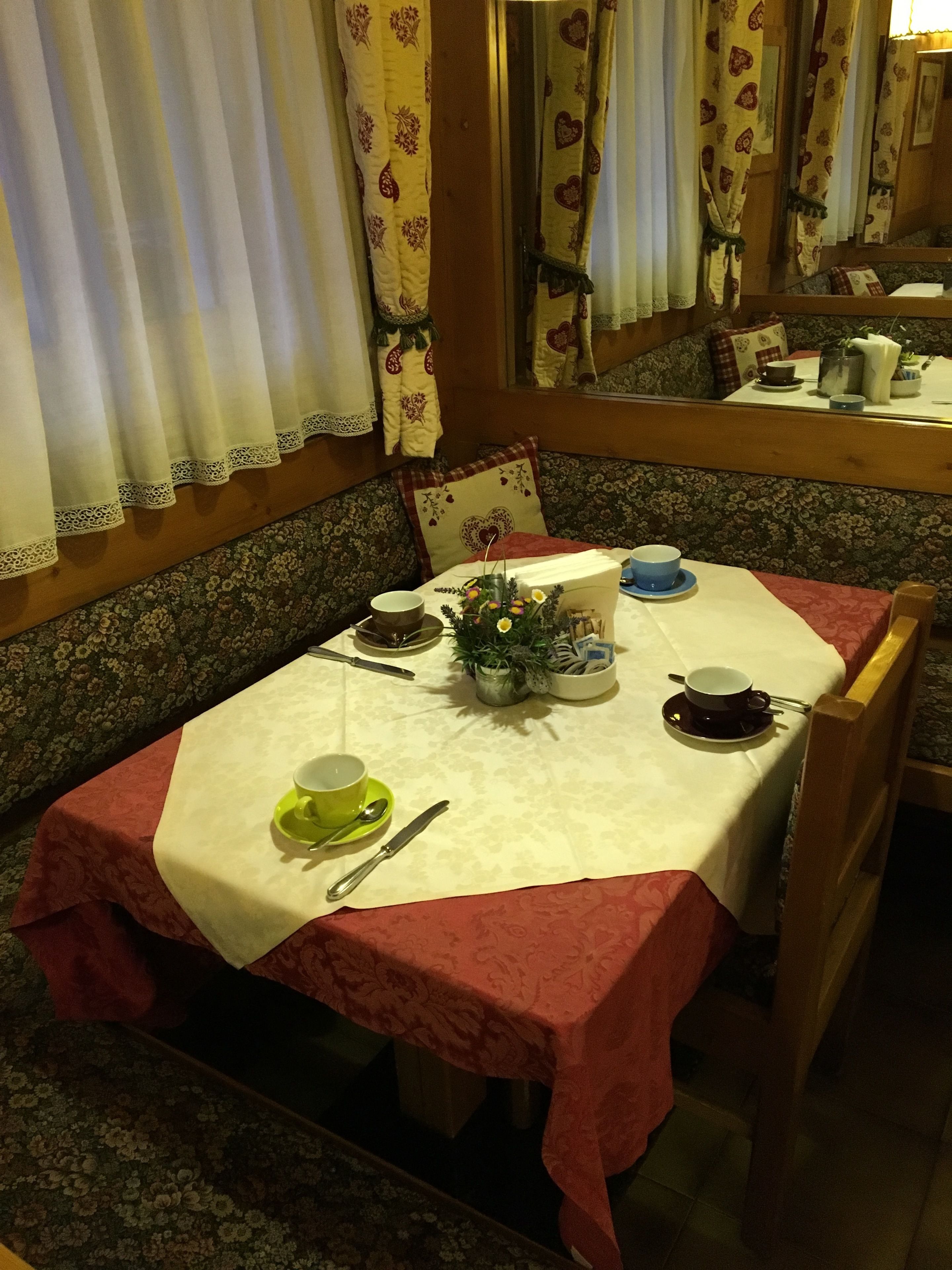 Un comedor con una mesa preparada para dos personas, con mantel floreado, un florero con flores y una variedad de vajilla.