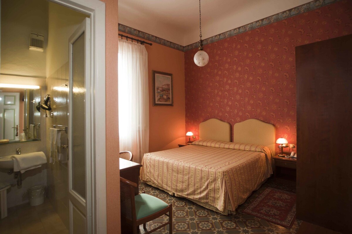 A hotel room with a bed, two bedside lamps, a mirror, a sink, and a chair.
