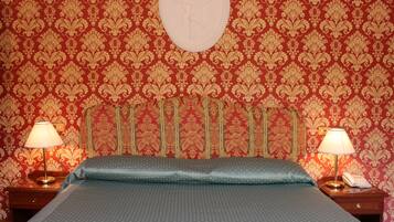 Une chambre d’hôtel avec un papier peint à motifs, un lit recouvert d’une courtepointe bleue, deux lampes de chevet et un miroir rond accroché au mur.