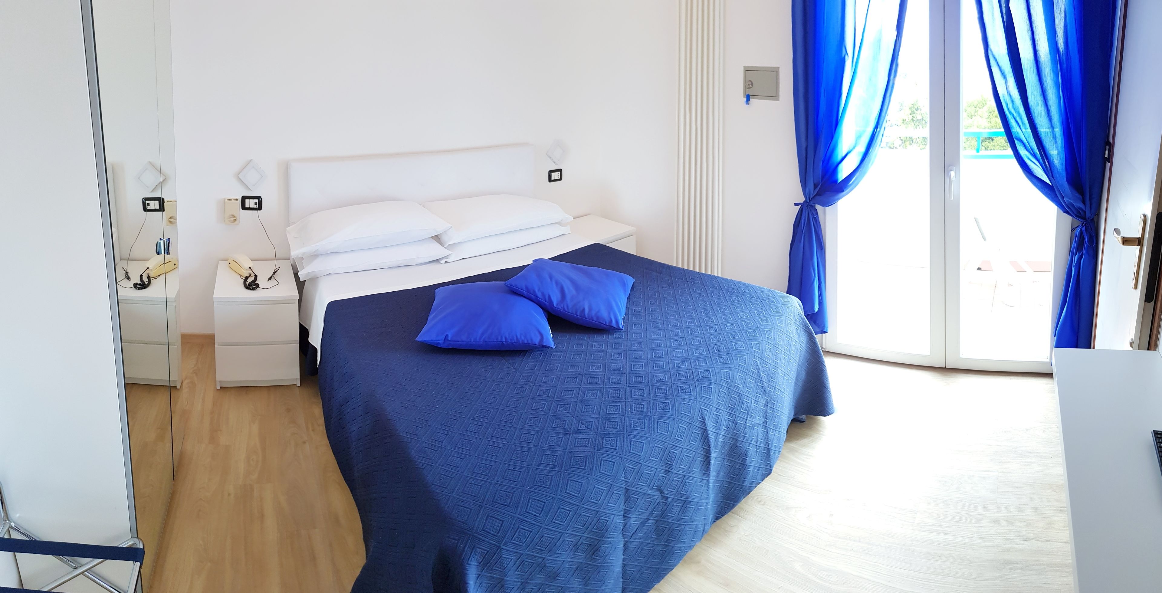 Uma cama bem arrumada com colcha azul e dois travesseiros, uma mesa de cabeceira com telefone e uma janela com cortinas azuis.