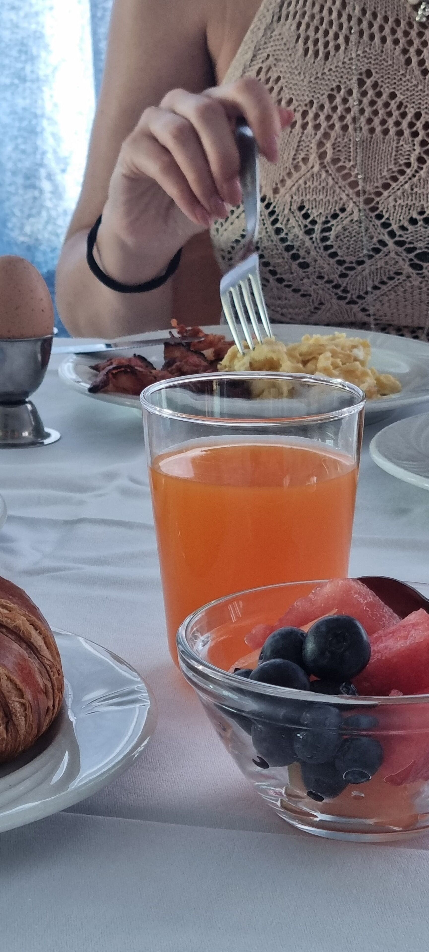 Une table mise pour le petit-déjeuner avec un croissant, un verre de jus d’orange et une assiette contenant des œufs et du bacon.