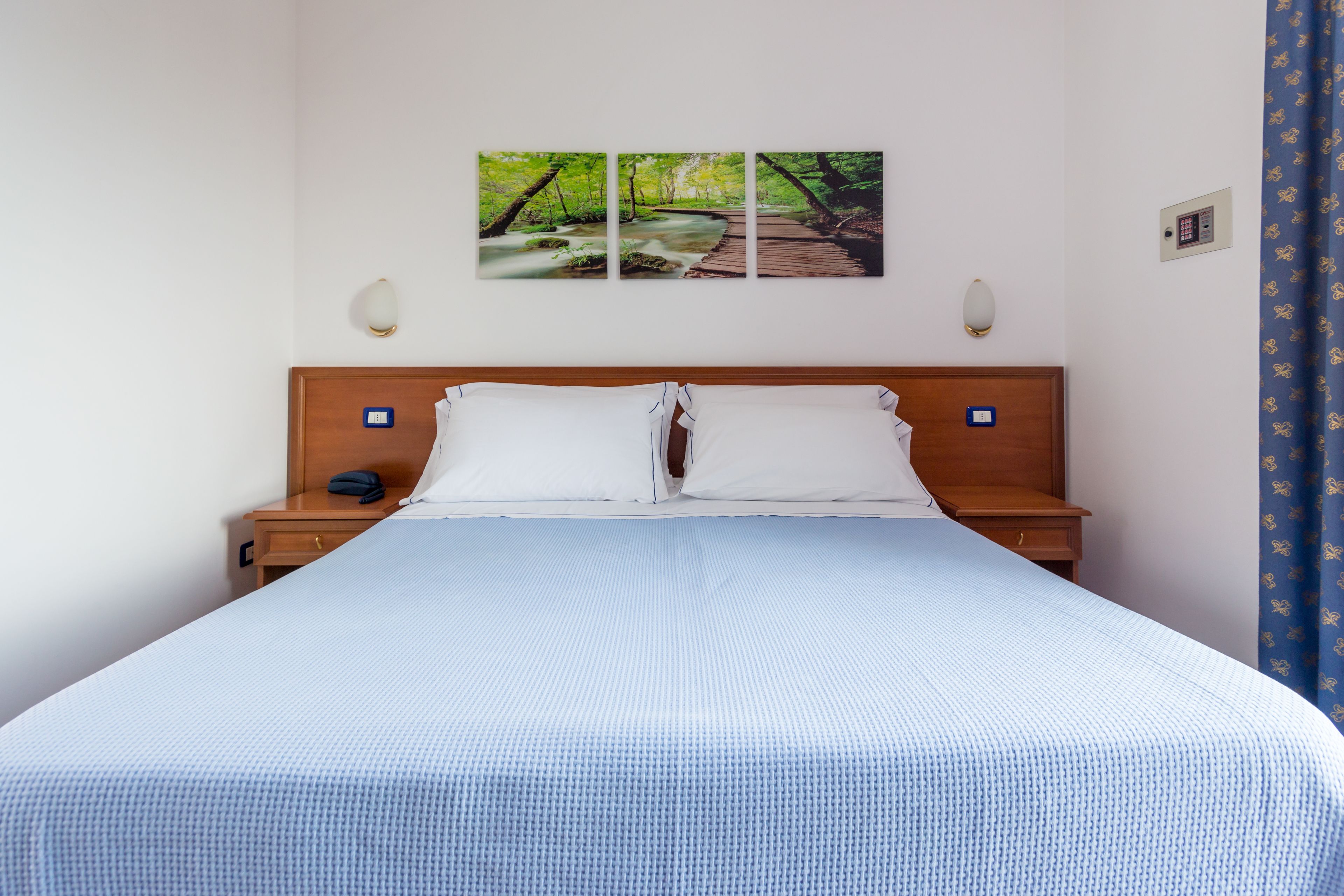 A hotel room with a large bed, a wooden headboard, two bedside tables, a nightstand, a wall-mounted lamp, and a painting of a bridge over a stream.