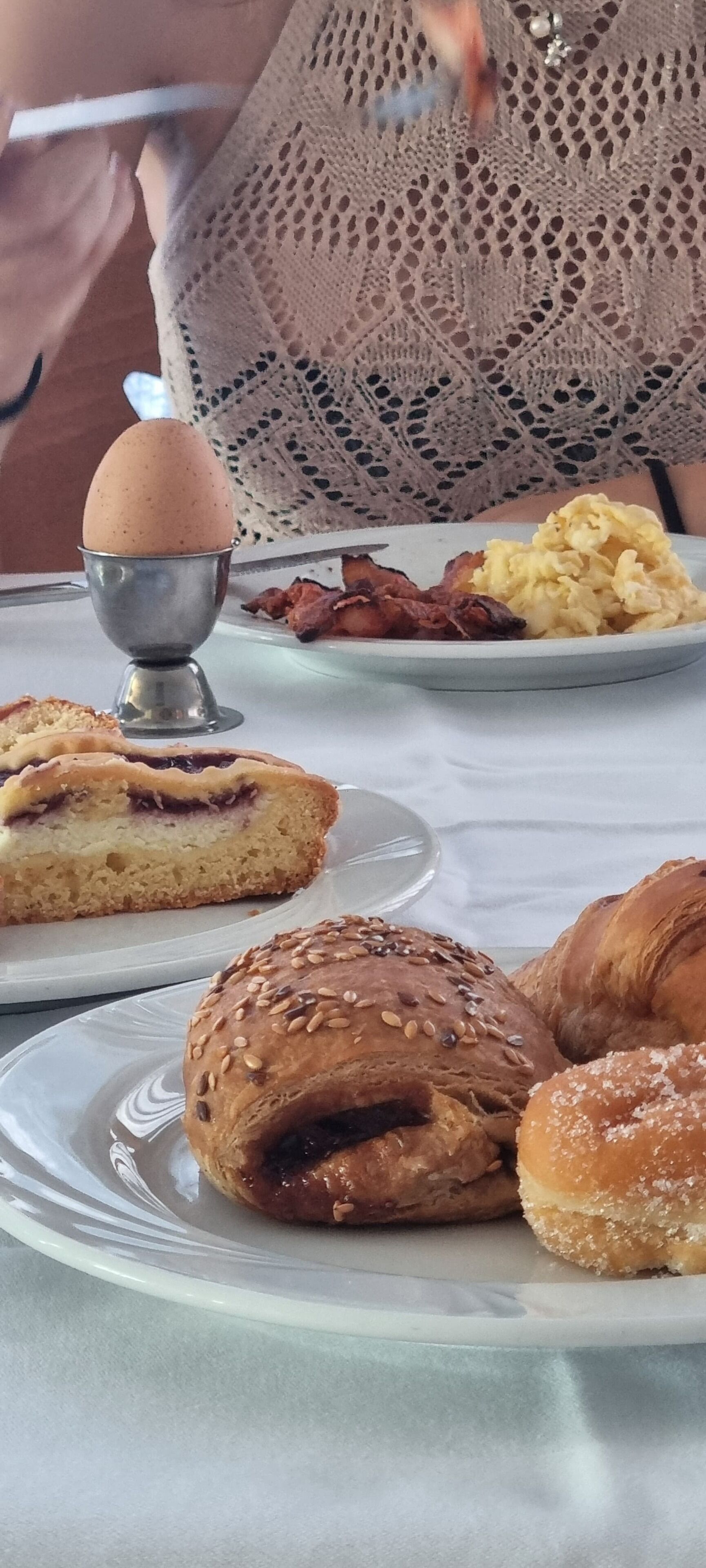 A breakfast table with various pastries, a metal egg cup, and a plate of bacon and scrambled eggs.