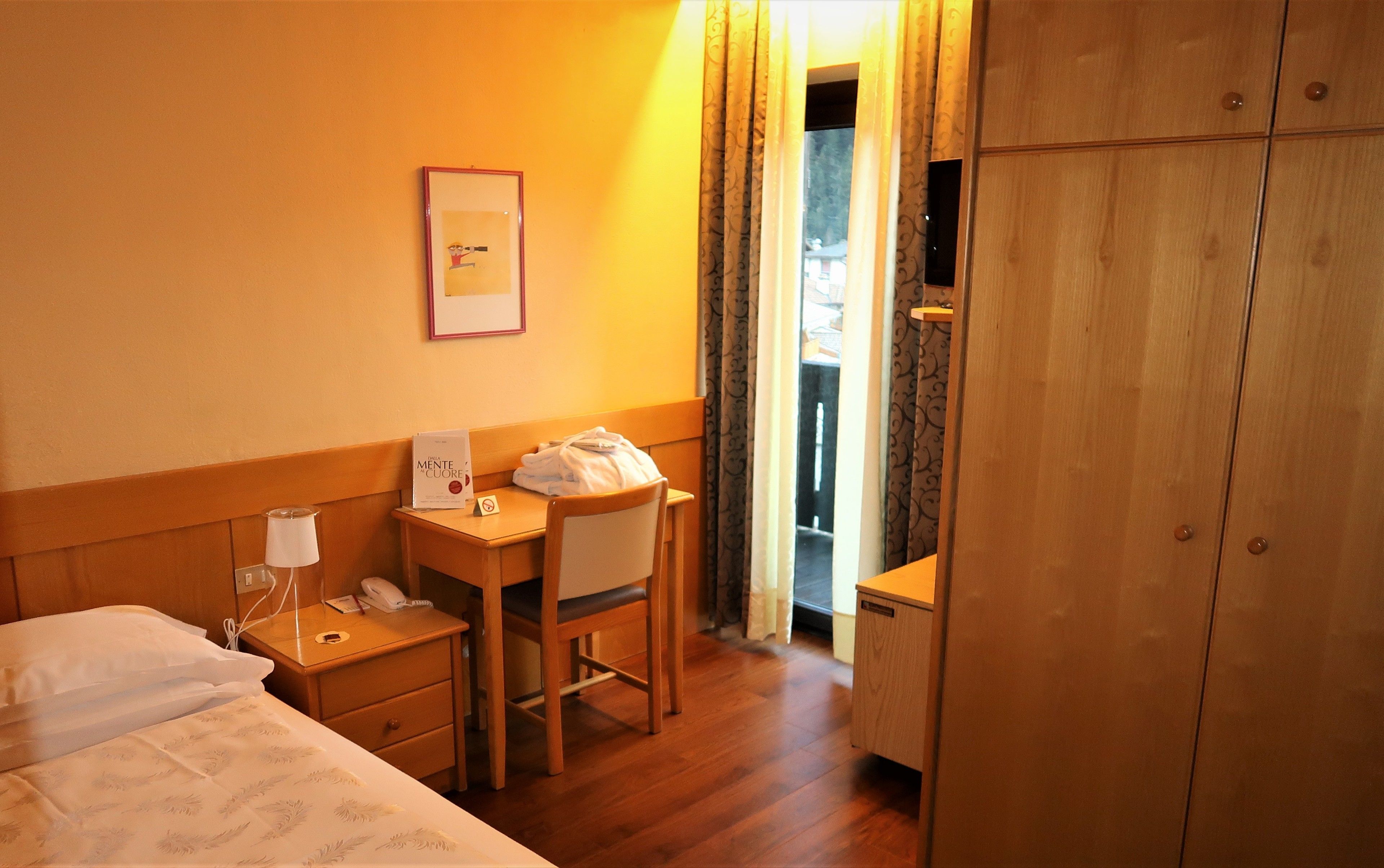 A hotel room with a bed, a desk with a chair, a telephone, a framed picture on the wall, and a wardrobe.