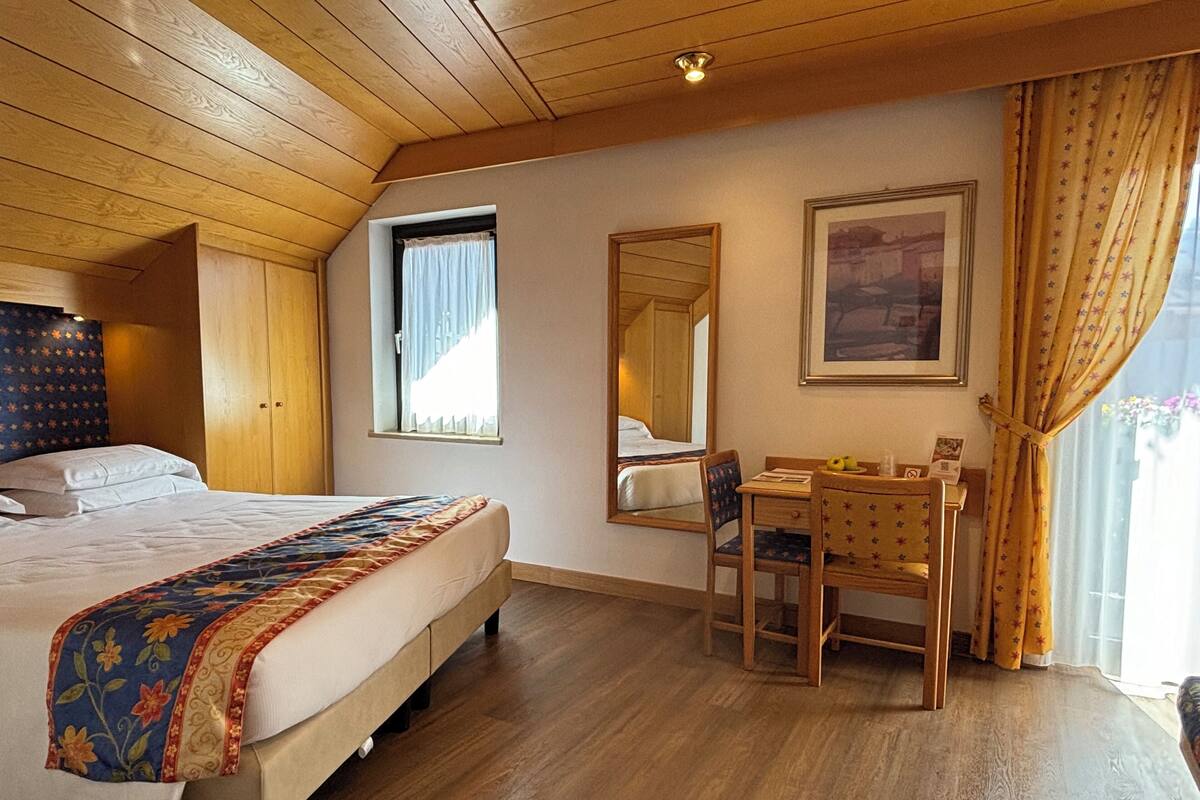 A hotel room with a bed, a desk, a chair, a window, a mirror, and a painting on the wall.