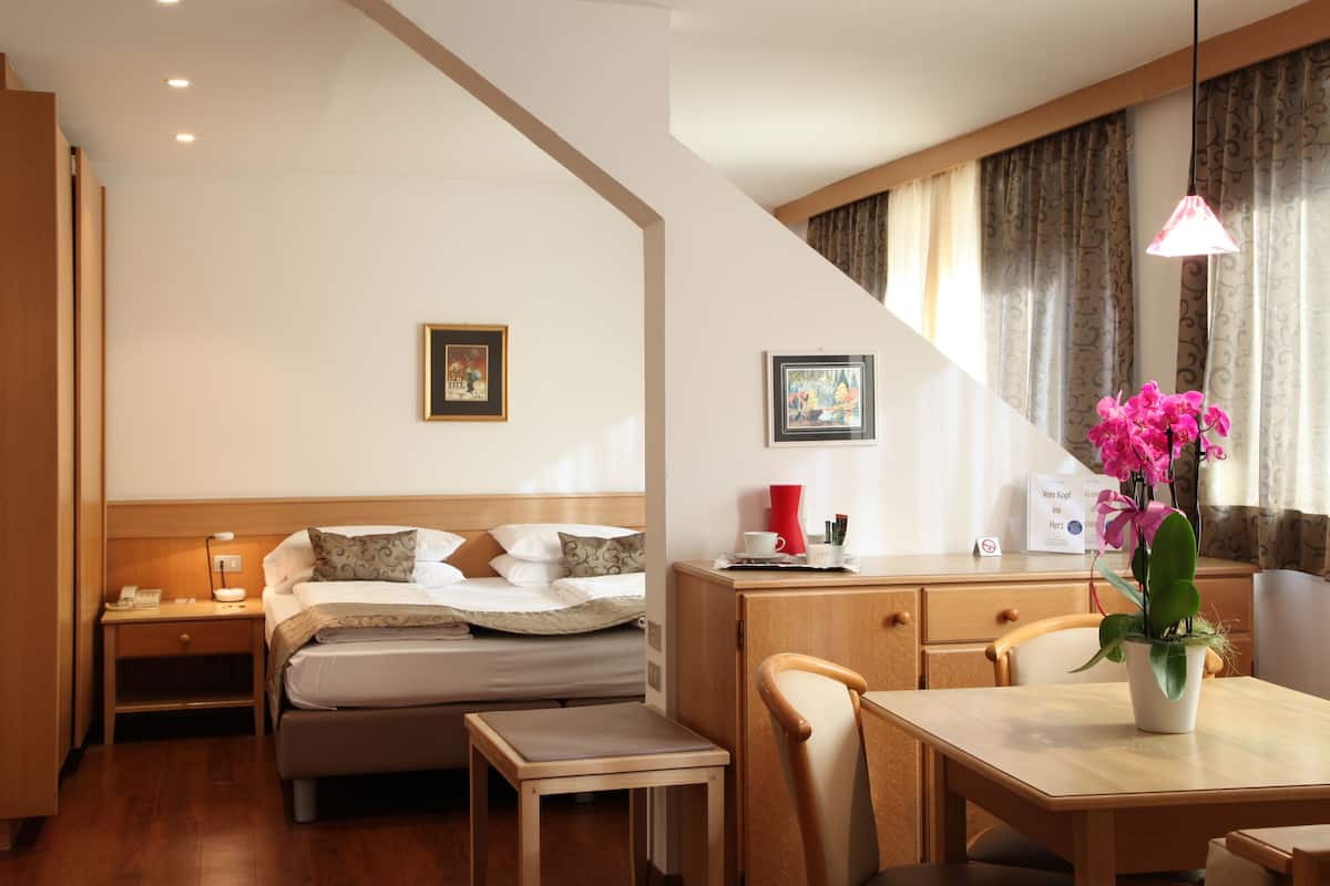 A hotel room with a bed, bedside table, desk, chair, and a dining table with a potted plant.