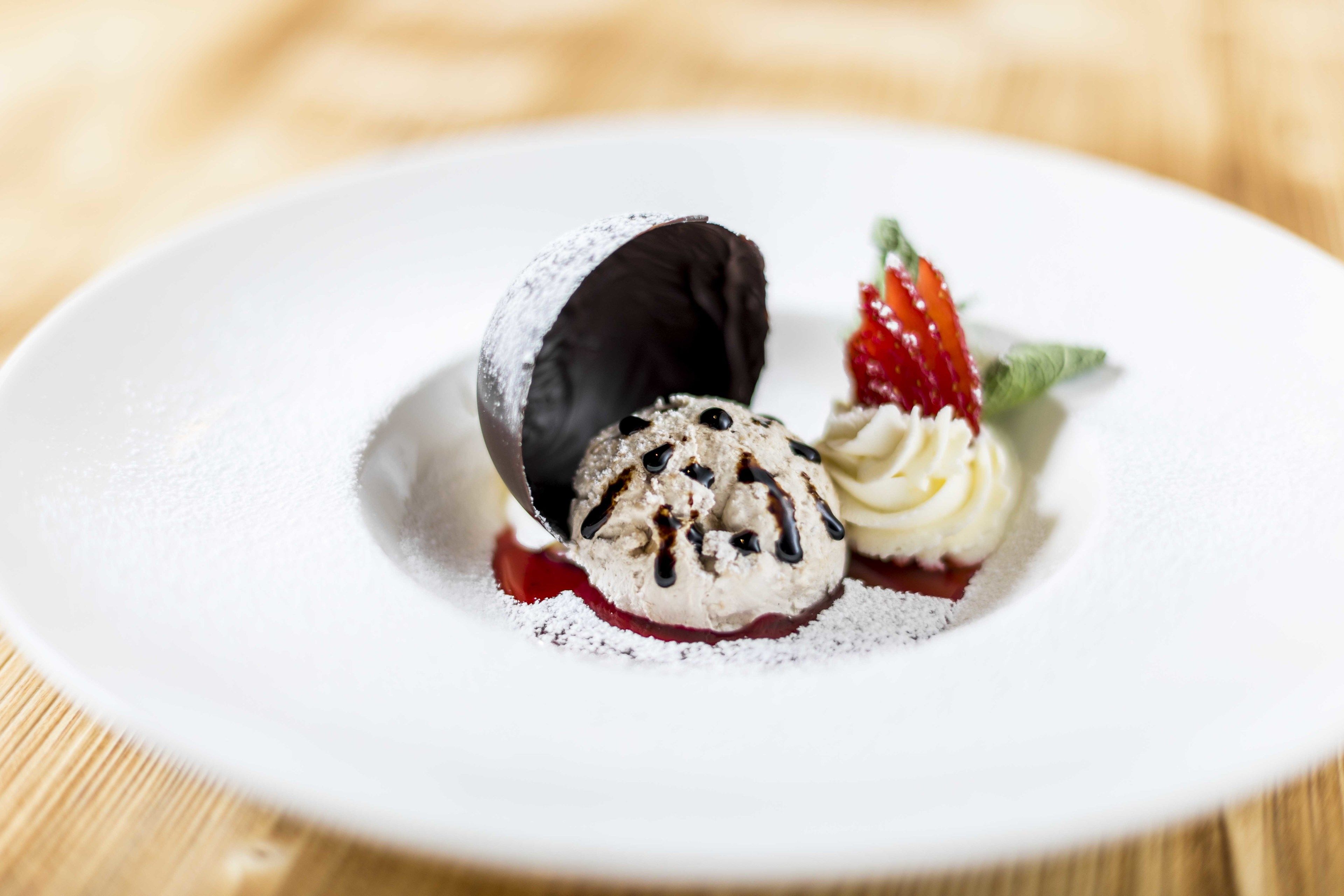 Un plato con postre, una cobertura de chocolate, una bola de helado con salsa de chocolate y una fresa sobre un plato blanco.