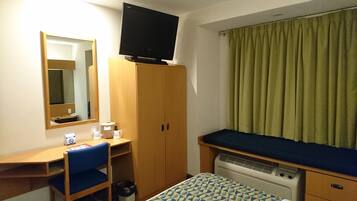 A small hotel room with a wooden desk, a chair, a bed with a blue mattress, a television on a stand, a heater, a mirror, and a window with green curtains.