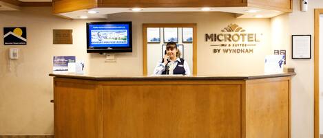 Una recepción en el Microtel Inn & Suites by Wyndham, con un mostrador de madera, un televisor montado y un cartel de bienvenida enmarcado.