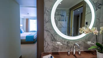 Une salle de bain d’hôtel avec un lavabo en marbre, un miroir rond et une vue sur une chambre à coucher avec un lit et une fenêtre.