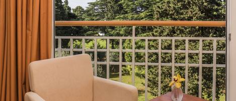 A room with a beige armchair, a wooden table, and a view of a green garden through a white grid window.