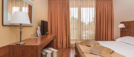 A hotel room with a wooden desk, a television, a bed with beige bedding, and a large window with curtains.