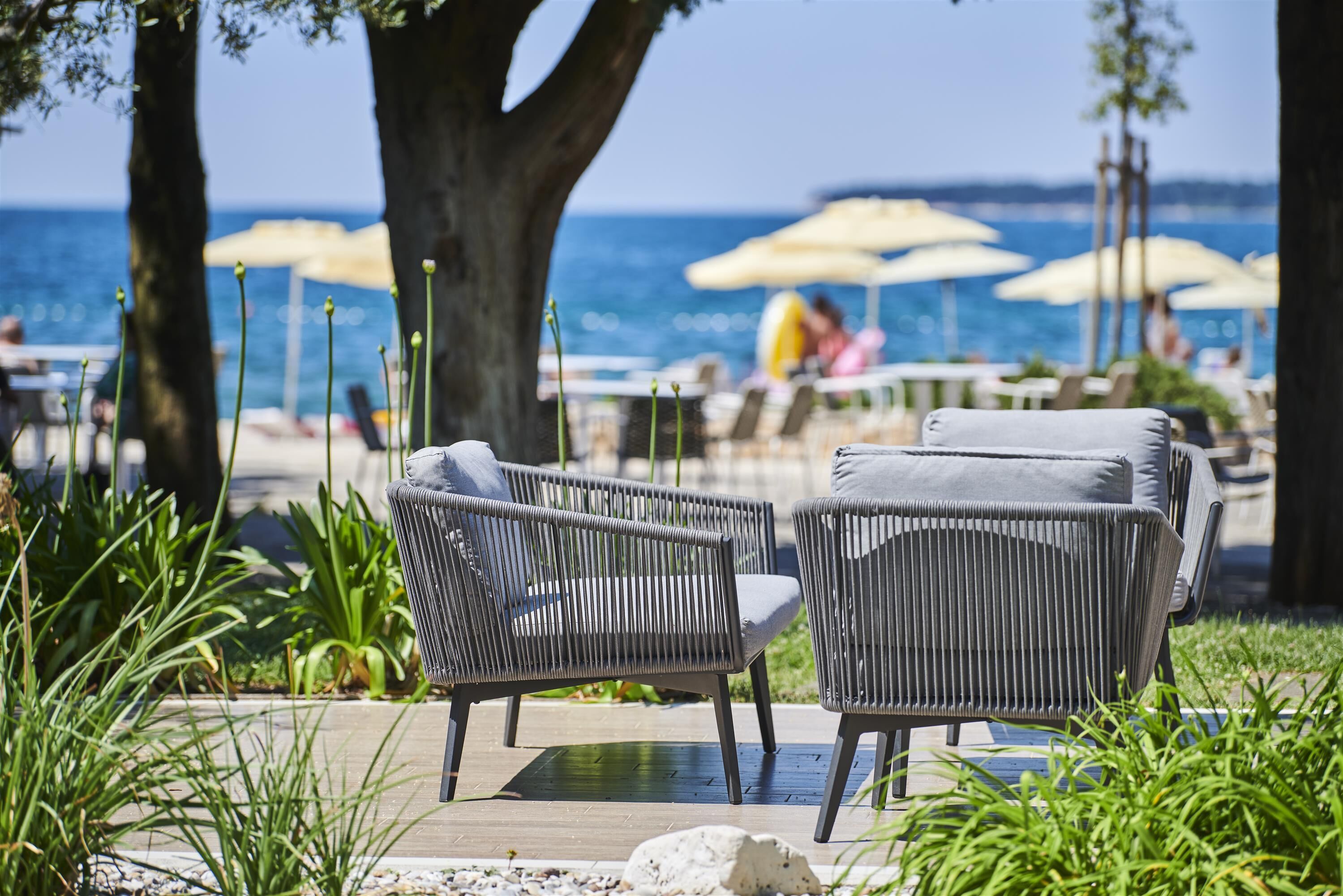 A patio area with wicker chairs and a table, overlooking a beach with umbrellas and people in the background.