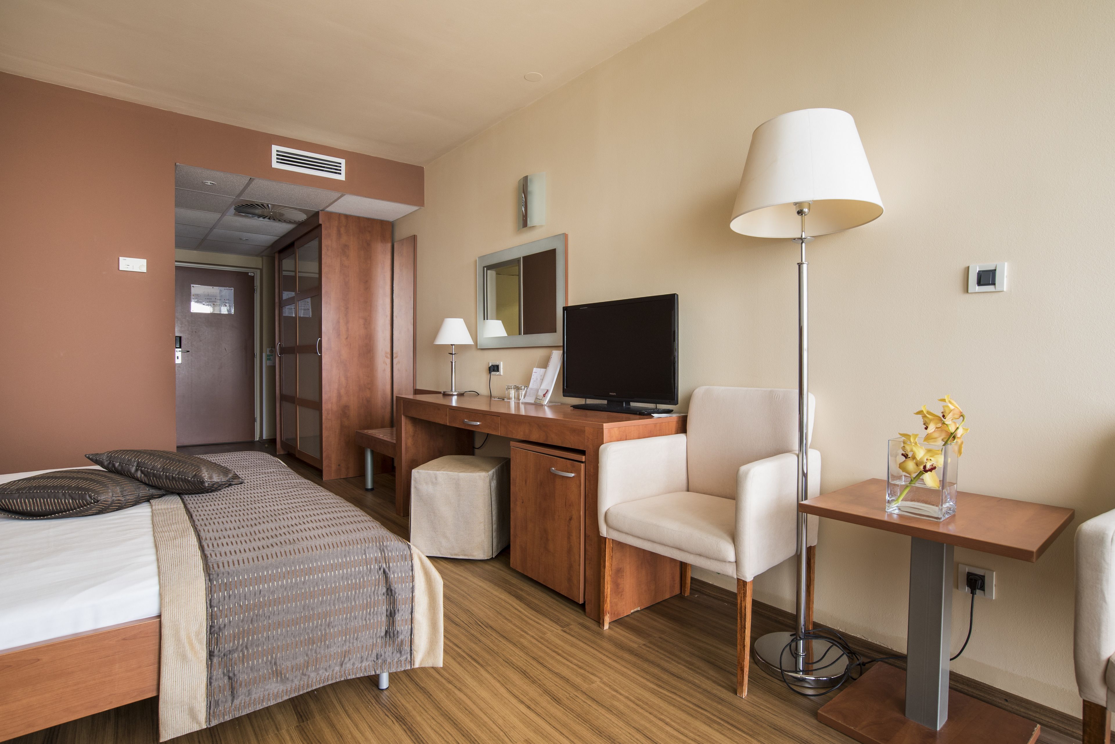 Habitación de hotel con cama, escritorio, silla, televisor, lámpara y una mesita con flores.
