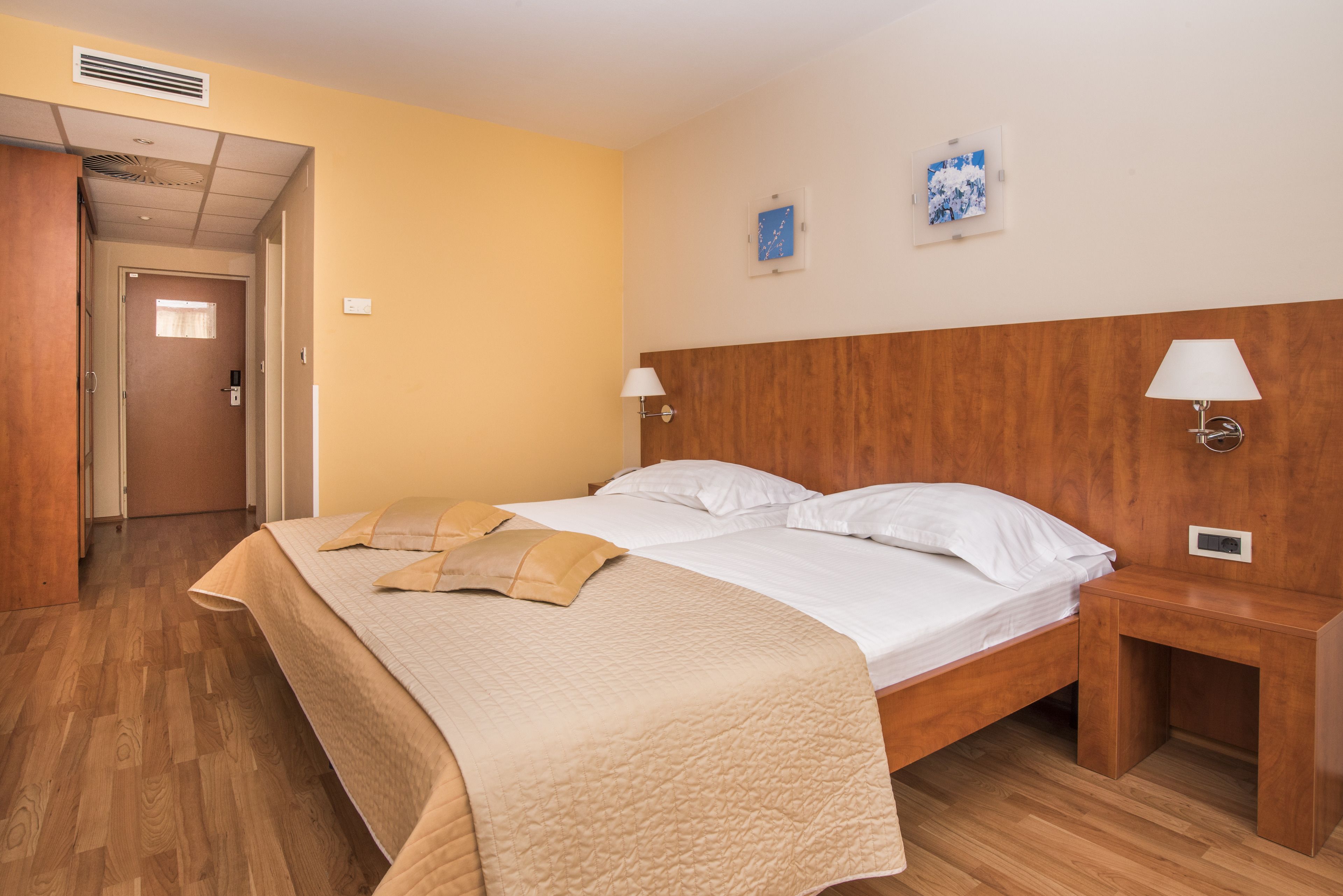 A hotel room with a wooden headboard, a bed with white linens, a beige quilt, two bedside lamps, and a wooden nightstand.