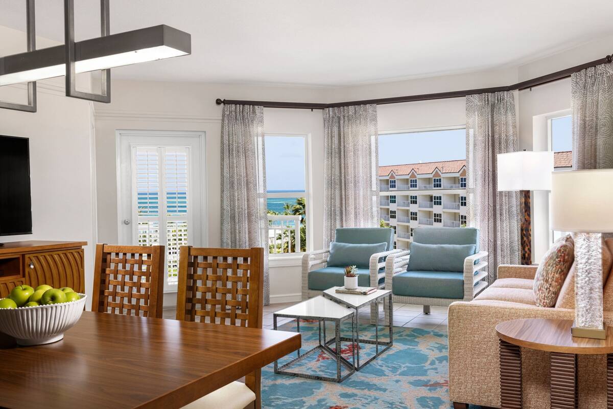 A modern living room with a wooden dining table, a bowl of green apples, and a view of the ocean through large windows.