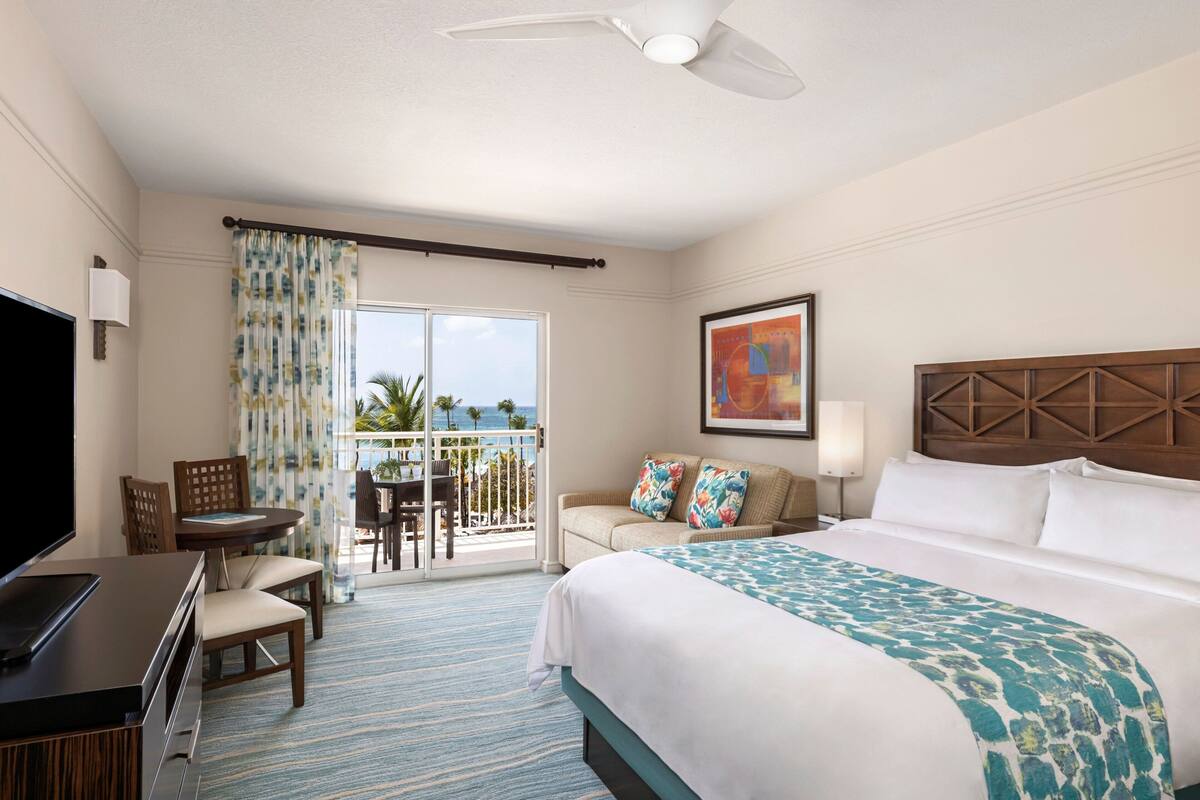 A hotel room with a large bed, a TV, a balcony with a view of the ocean, and a dining area with a table and chairs.