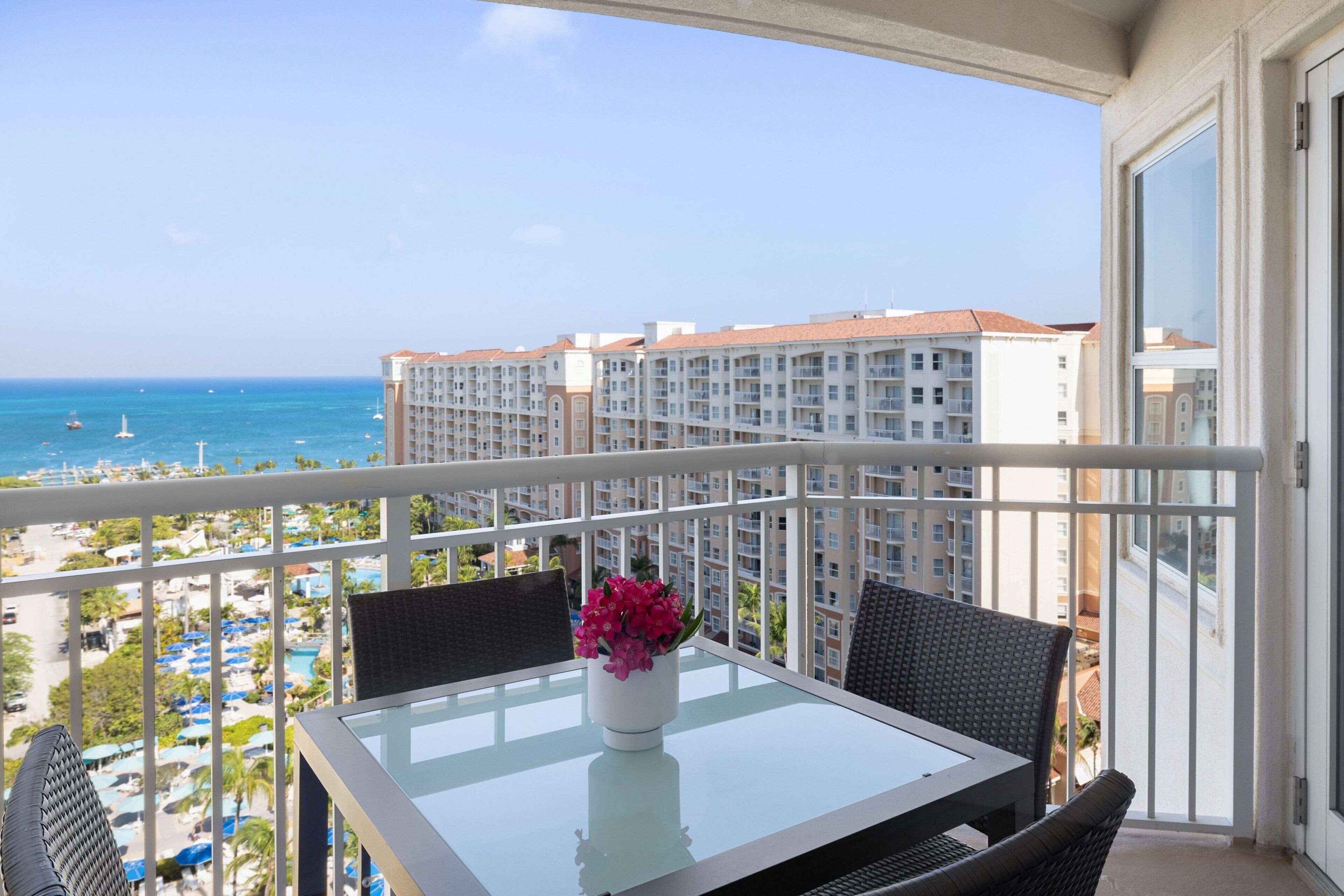 A balcony with a glass table and chairs, overlooking a beach and hotel buildings.