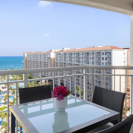 A balcony with a glass table and chairs, overlooking a beach and hotel buildings.