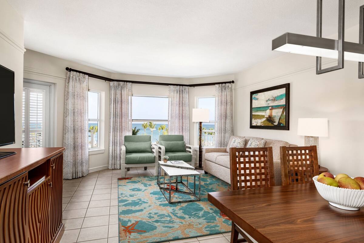 A living room with a TV, seating, a dining table, and a view of the ocean.