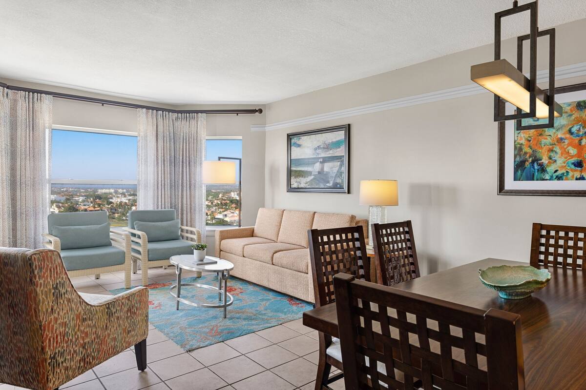 A hotel room with a balcony, a sofa, a dining table, and chairs.