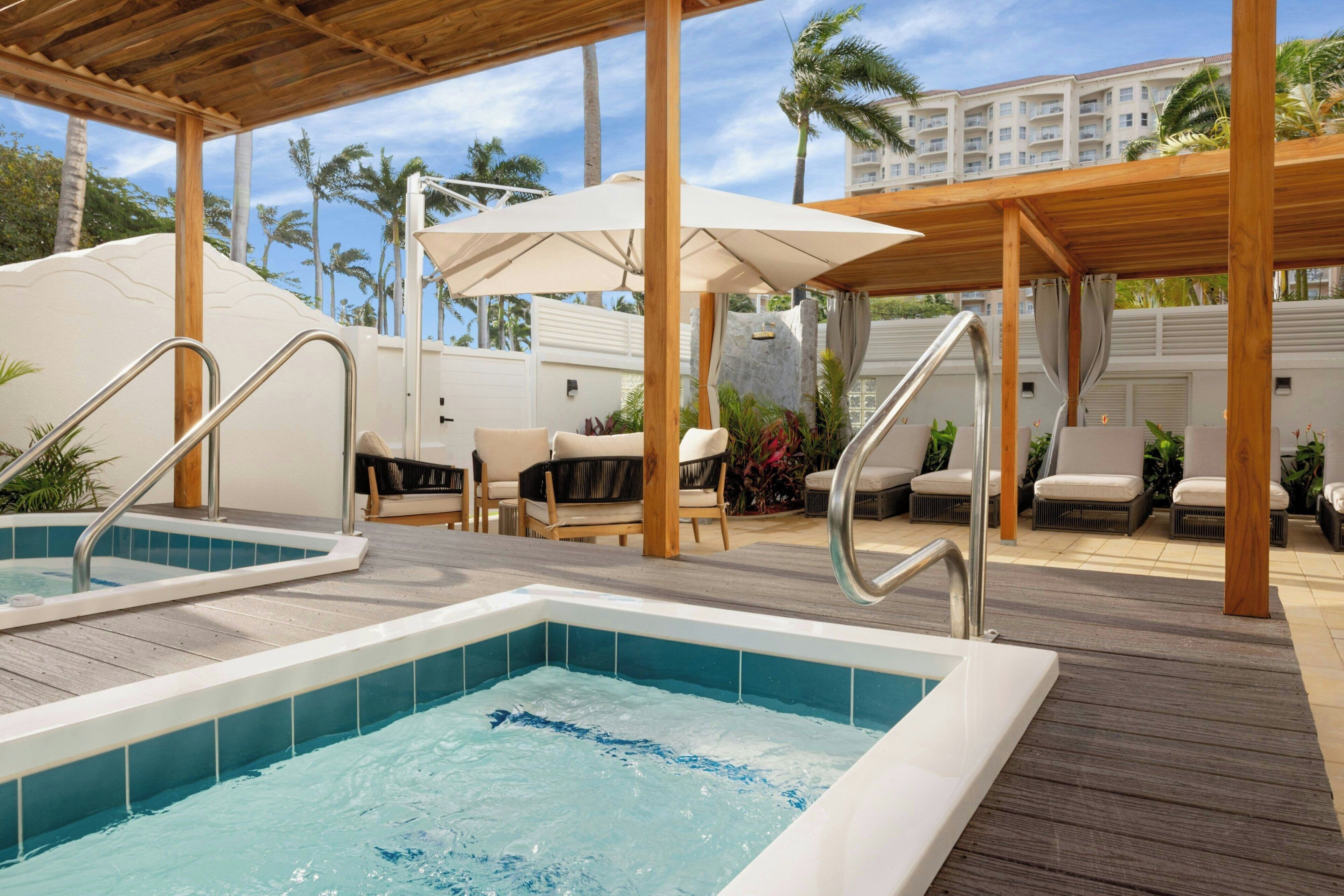 Un espace piscine comprenant deux bains à remous, une terrasse en bois, des chaises longues et des parasols.