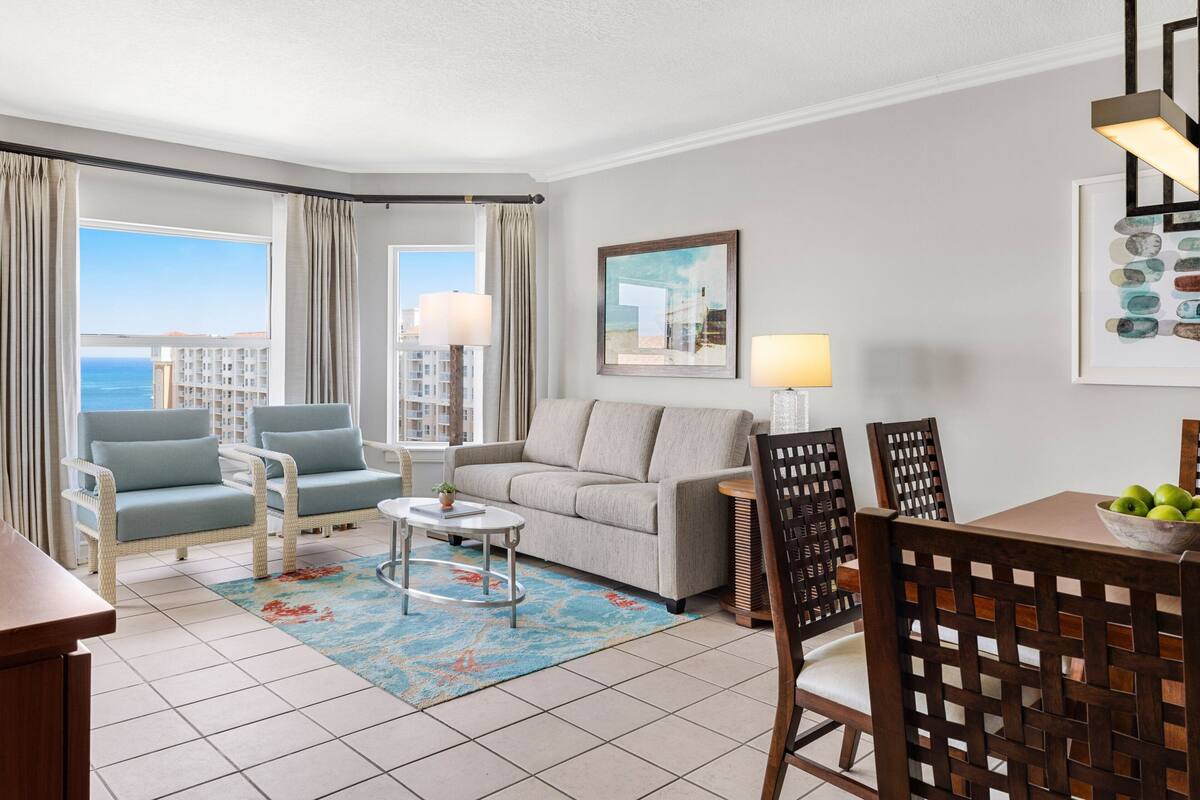 A hotel room with a balcony, a sofa, a dining table with chairs, and a view of the ocean.