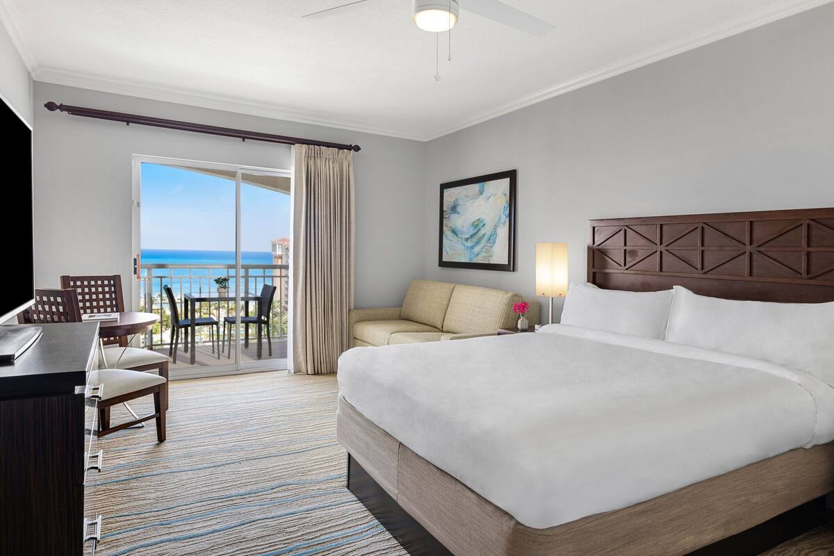 A hotel room with a bed, a desk, a chair, a balcony with a view of the ocean, and a painting on the wall.