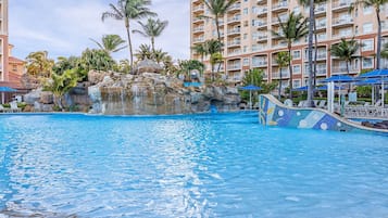 Una piscina con una formación rocosa y palmeras, rodeada por un edificio de hotel de varios pisos.