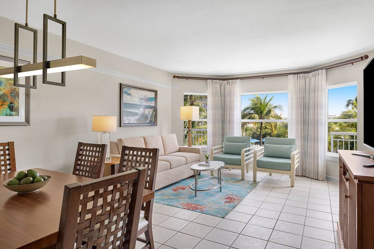 A living room with a sofa, chairs, a dining table, and a TV. There are windows with curtains and a view of palm trees.