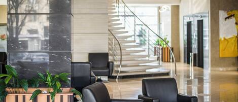 A modern lobby with a marble reception desk, black leather seating, a spiral staircase, and a glass door leading to a hallway.