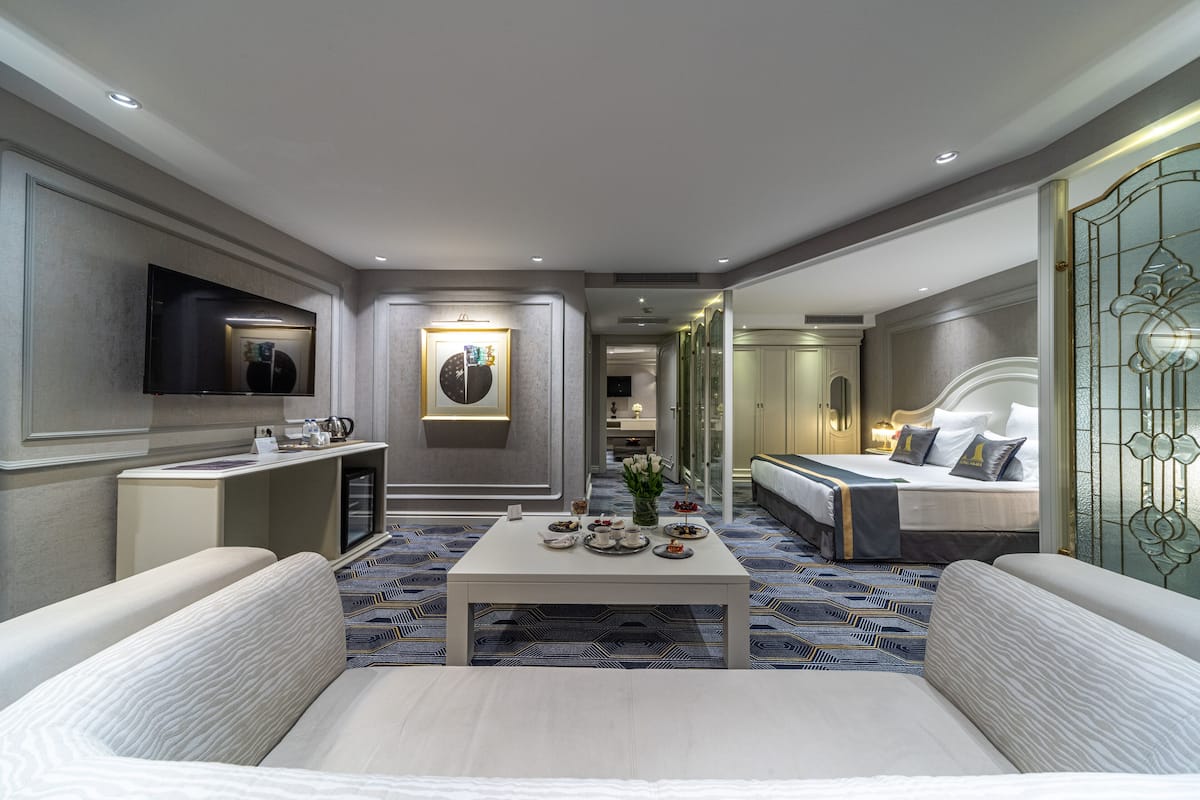 Ein modernes Hotelzimmer mit einem großen Bett, einer Couch, einem Couchtisch mit Teeservice, einem flachbildfernseher an der Wand und einem dekorativen Spiegel.