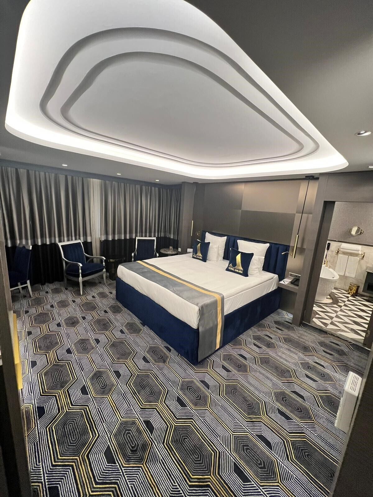 A modern hotel room with a large bed, a seating area with chairs, a bathroom, and a geometric patterned floor.