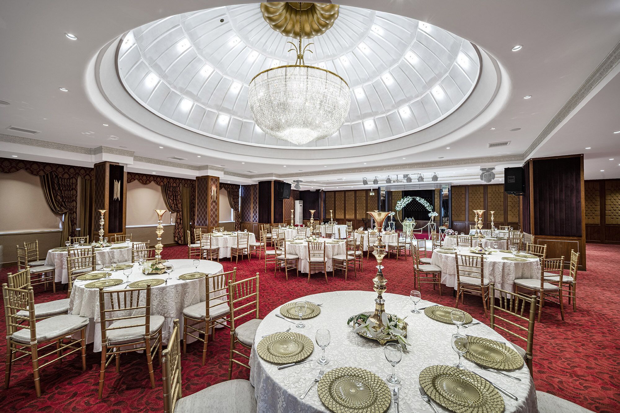 Een balzaal met ronde tafels, gouden stoelen en een centrale kroonluchter.