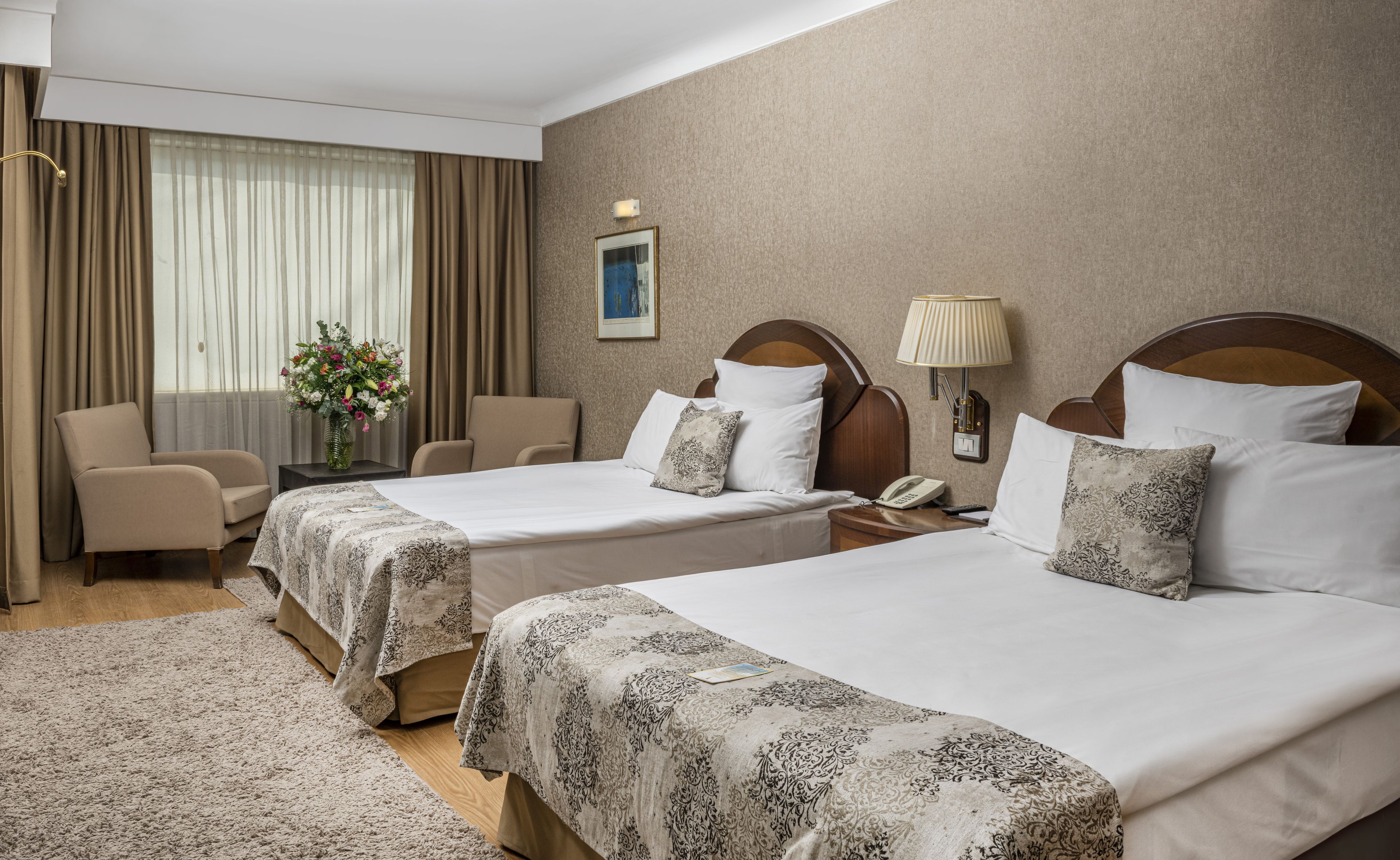 A hotel room with two beds, a desk, a chair, a lamp, and a vase of flowers.