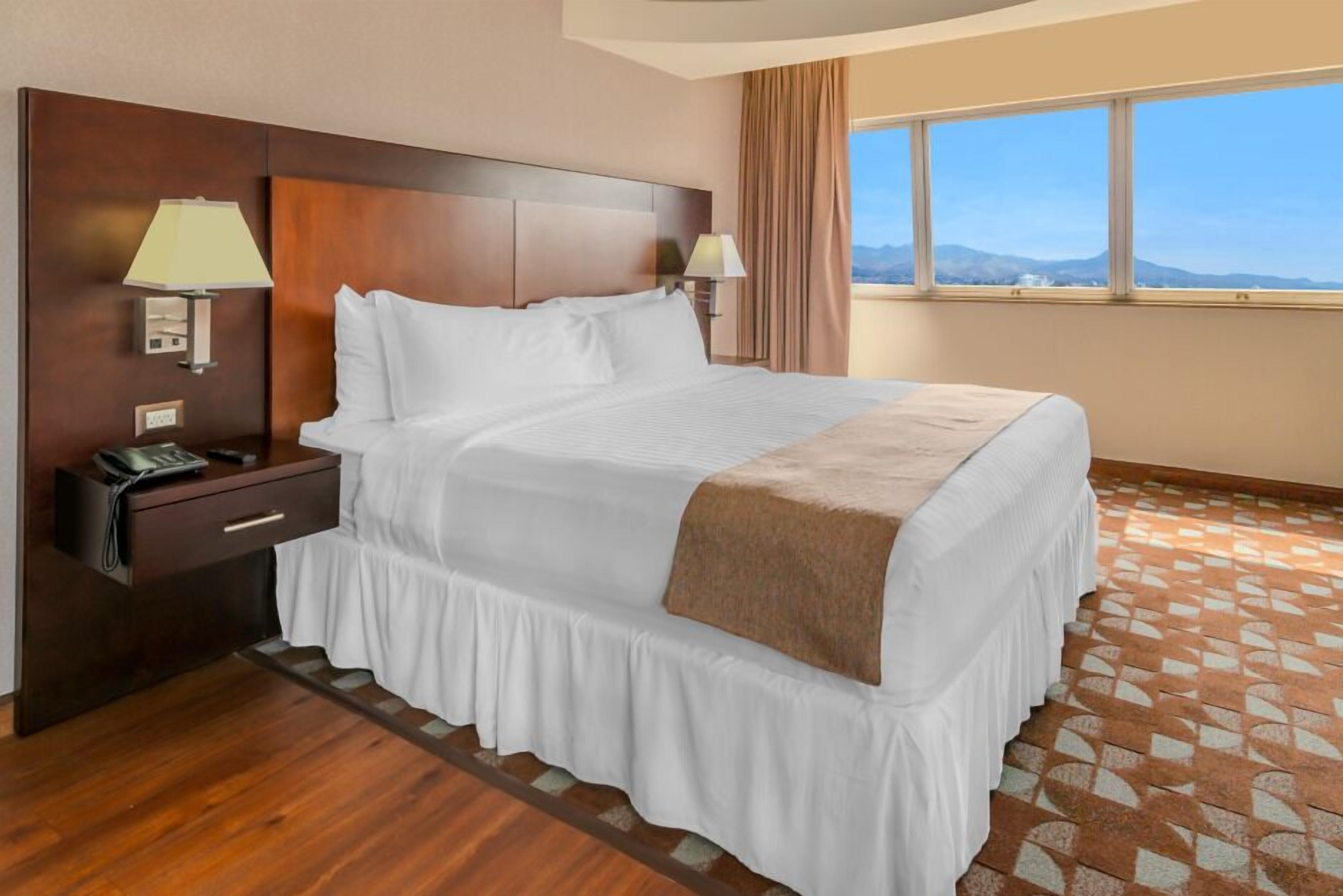 Habitación de hotel con una cama grande, dos lámparas de noche, una mesita de noche con un teléfono y una ventana con vistas a las montañas.
