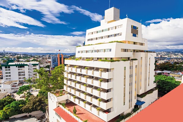 A multi-story building with balconies, surrounded by greenery and other buildings.